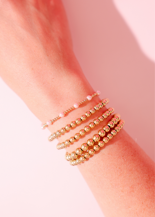 Gold beaded bracelets on a wrist against a pink background