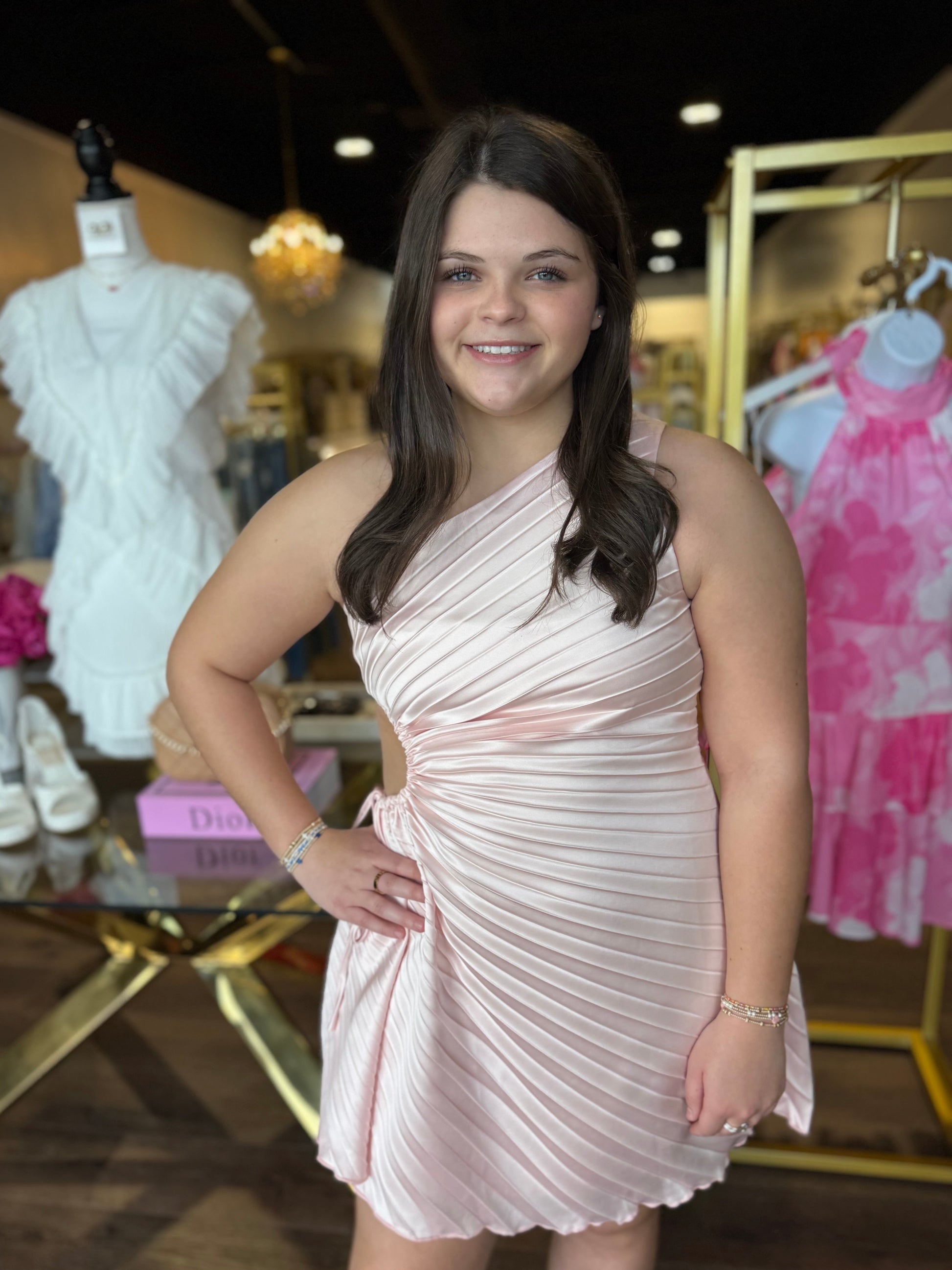 A young woman posing in a boutique wearing a light pink pleated one-shoulder mini dress. The background shows other clothing items on mannequins and racks inside a retail store.
