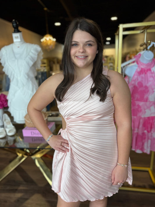 A young woman posing in a boutique wearing a light pink pleated one-shoulder mini dress. The background shows other clothing items on mannequins and racks inside a retail store.