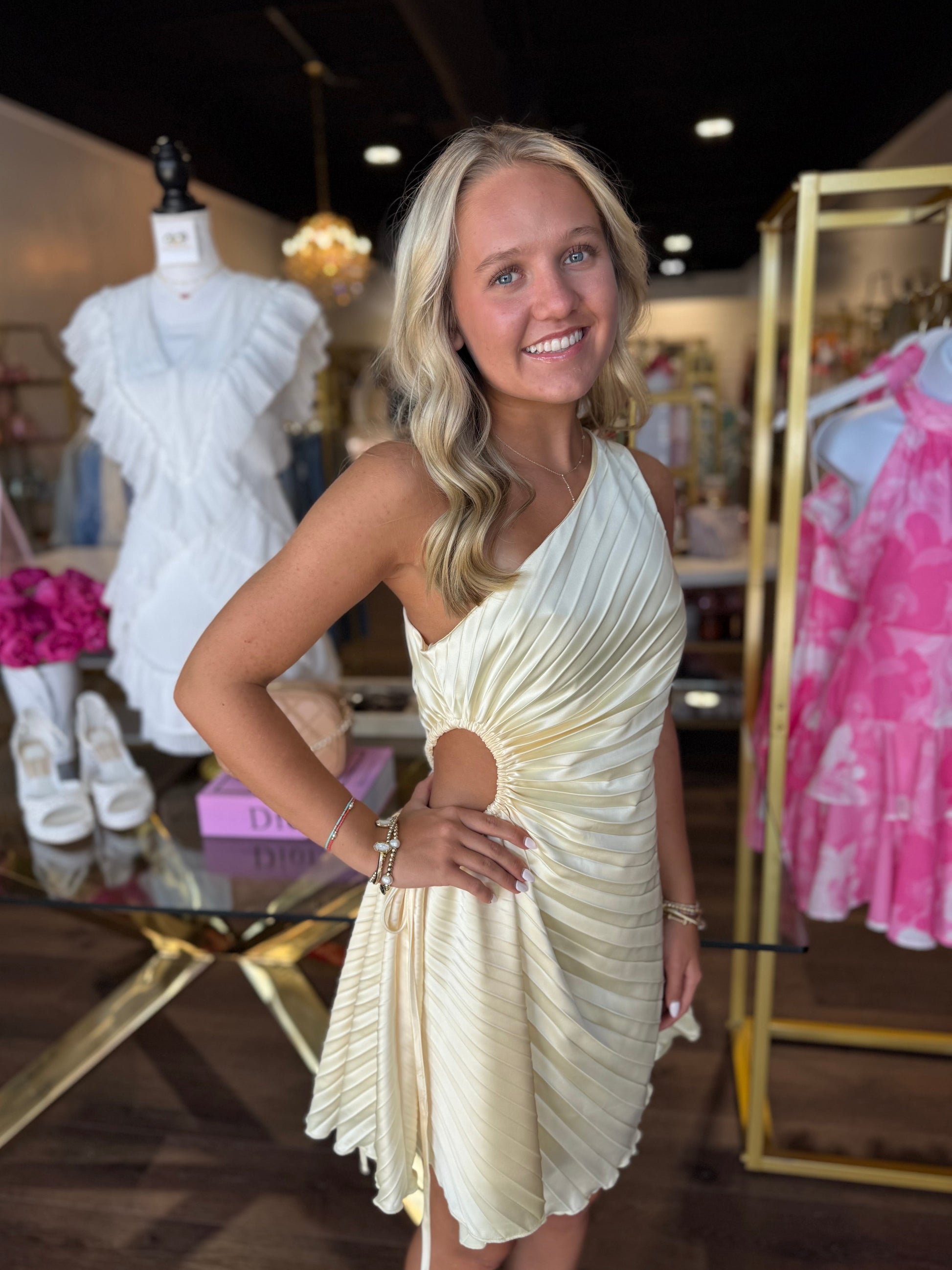 A smiling blonde woman wearing a pleated, one-shoulder cream dress with a side cutout, posing inside a boutique store with clothing racks and mannequins in the background.