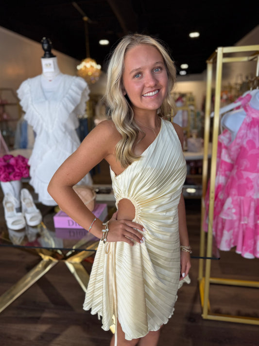 A smiling blonde woman wearing a pleated, one-shoulder cream dress with a side cutout, posing inside a boutique store with clothing racks and mannequins in the background.