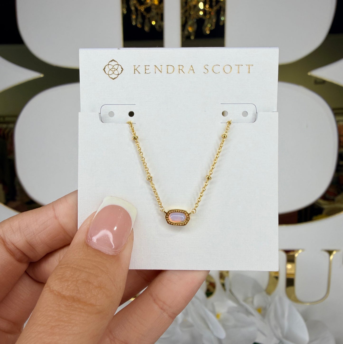 Gold necklace on a Kendra Scott card held by a hand with a blurred floral background