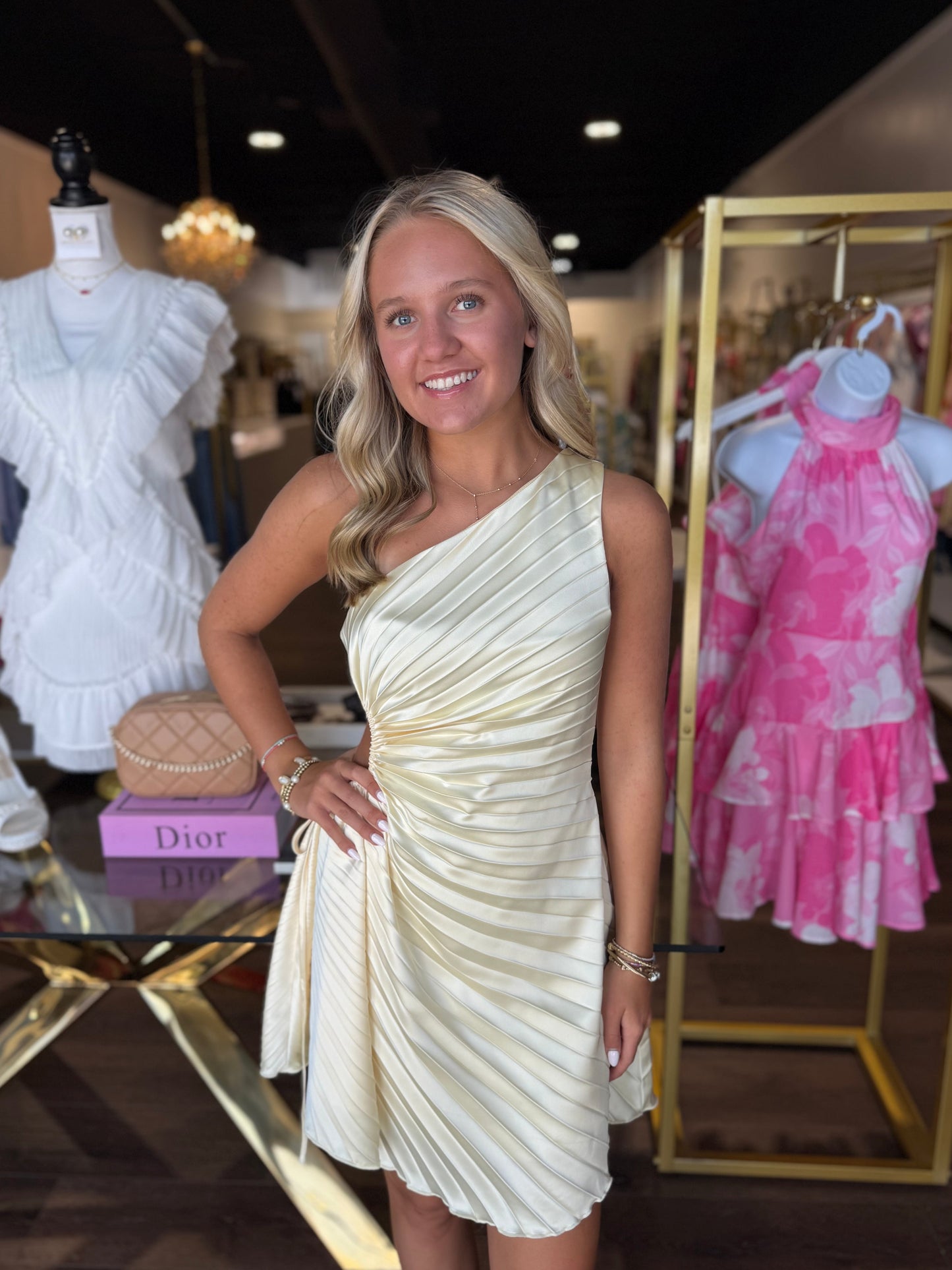 A smiling blonde woman stands in a boutique wearing a cream one-shoulder pleated dress. Mannequins display white and pink dresses in the shop, with a Dior box visible on a table.