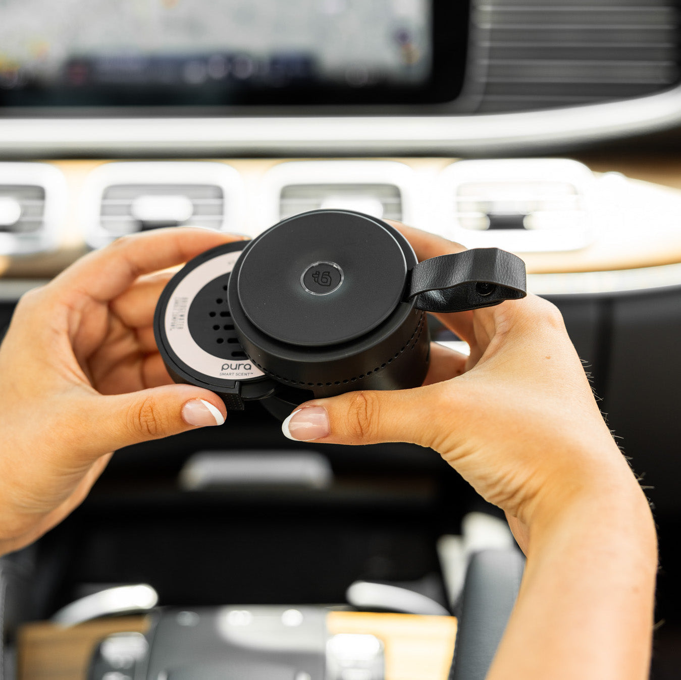 Person holding a black car air freshener with 'Pura' branding in a car interior.