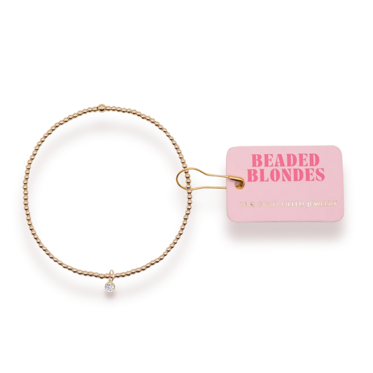 April Birthstone Charm Bracelet with a clear charm and gold beads beside a pink Beaded Blondes tag.