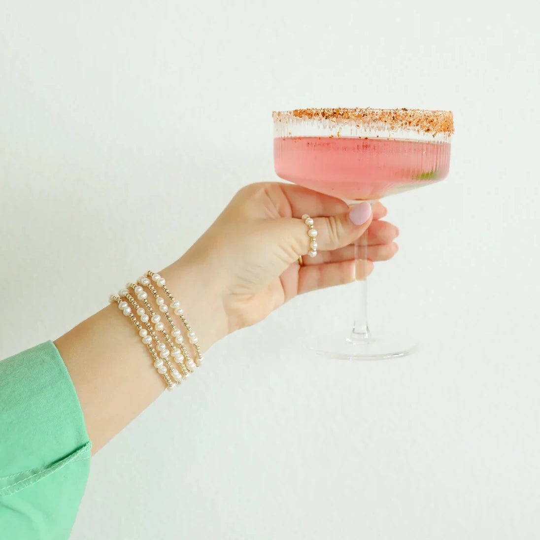 Pearl and gold bracelet stacks worn on a wrist while holding a pink cocktail glass.
