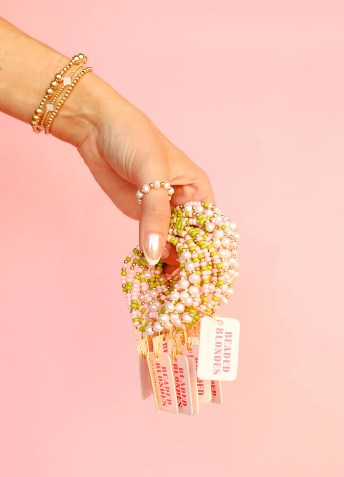 Pearl rings and gold bracelets styled on a hand holding strands of beaded jewelry against a pink backdrop.