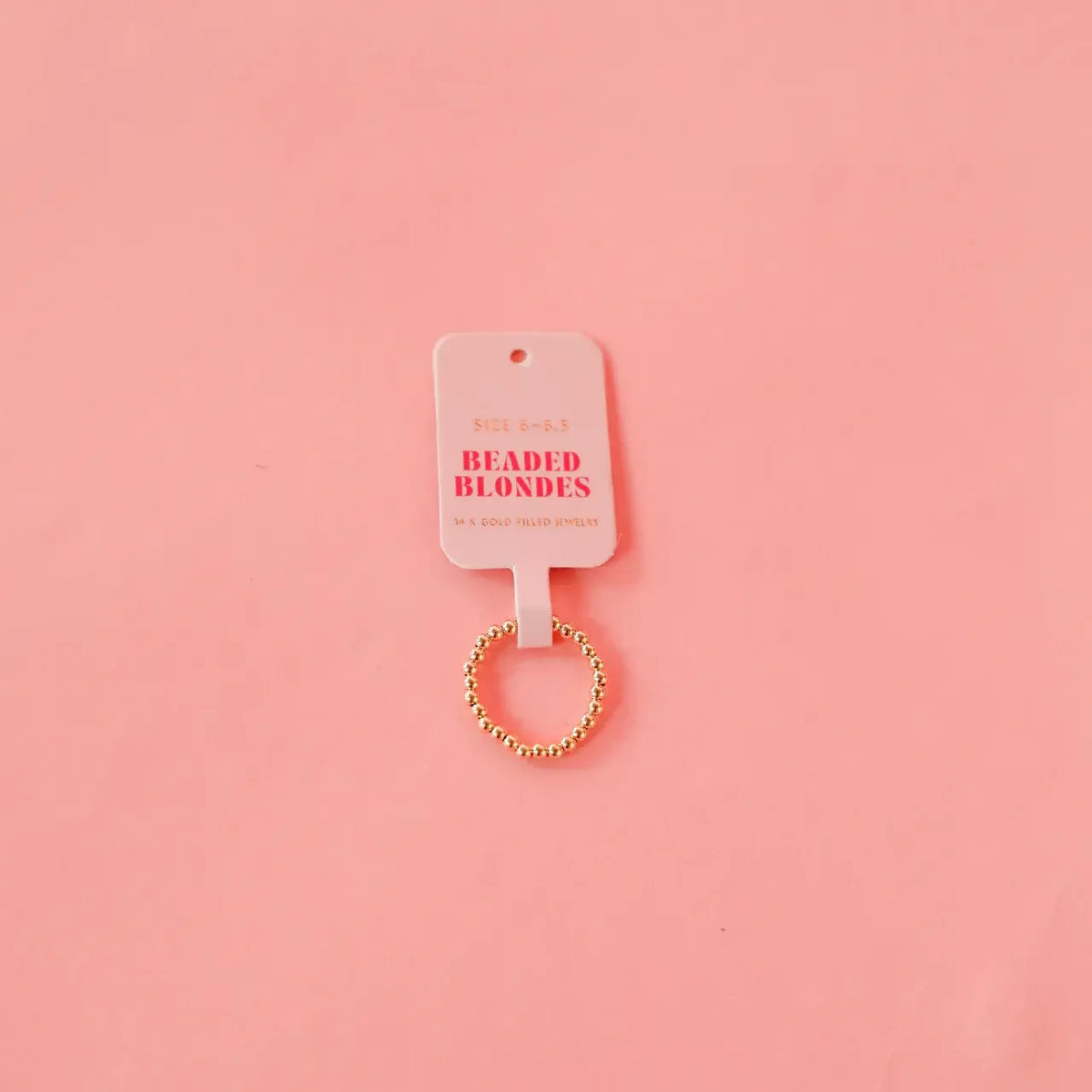 Ella gold beaded ring with a pink Beaded Blondes tag on a pink background.