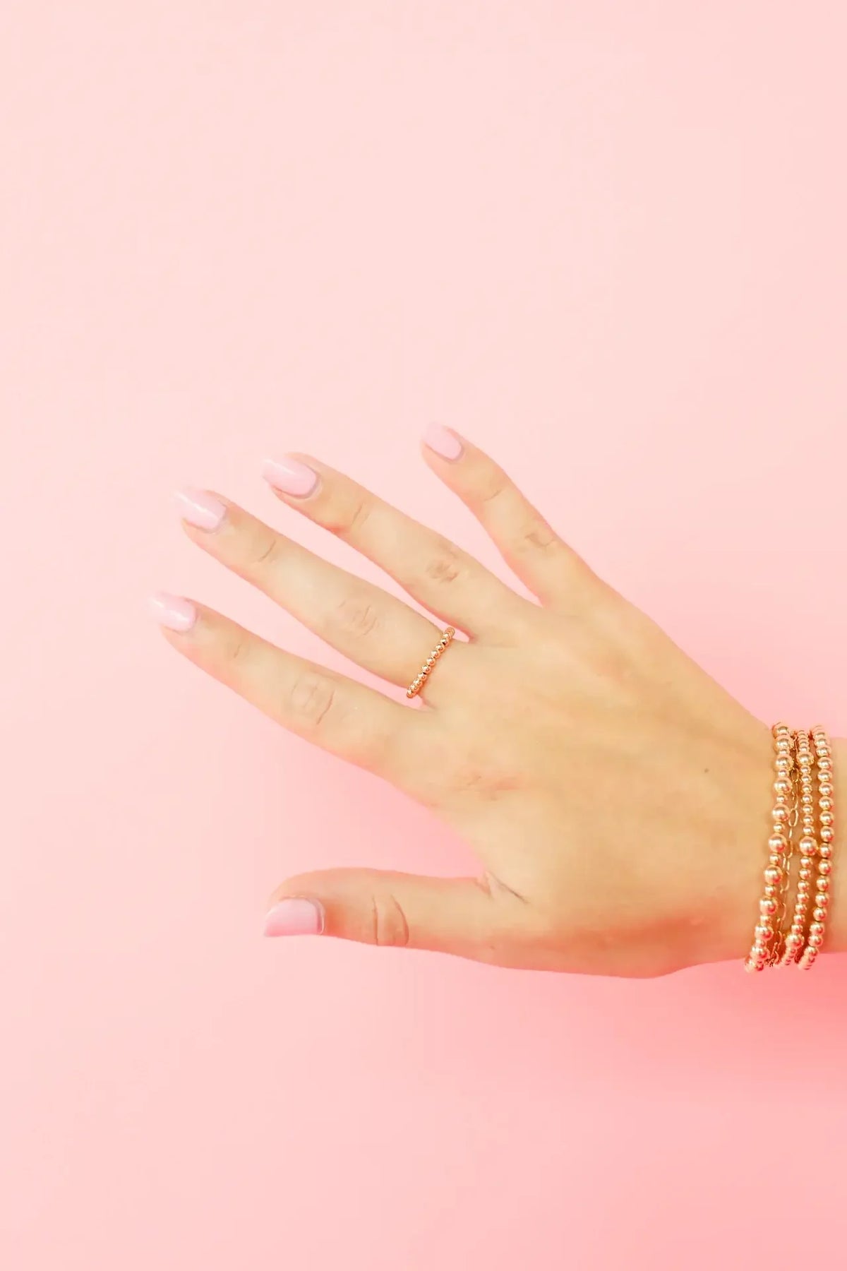 Gold beaded rings and layered gold bracelets worn together against a pink background.