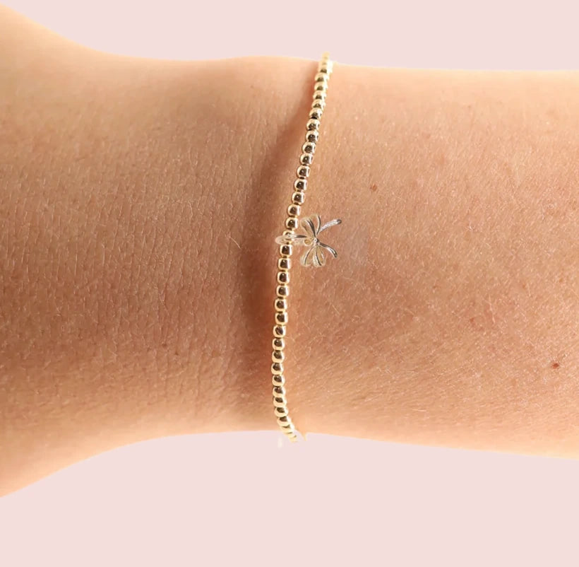 A delicate gold beaded bracelet with a bow charm worn on a wrist against a soft pink background.