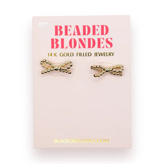 Beaded Blondes Gold Bow Earrings displayed as a pair on a pink jewelry card.