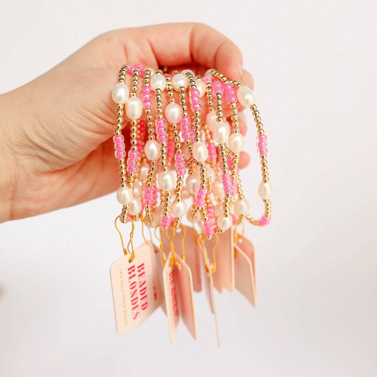 A hand holds several pink, pearl and gold beaded bracelets with pink display tags.