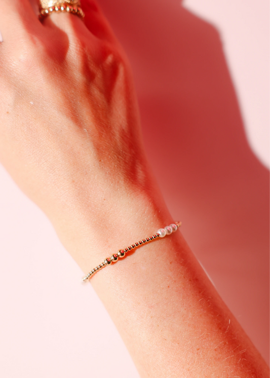 A delicate gold and pearl alternating bracelet worn alone on a wrist against a pink background.