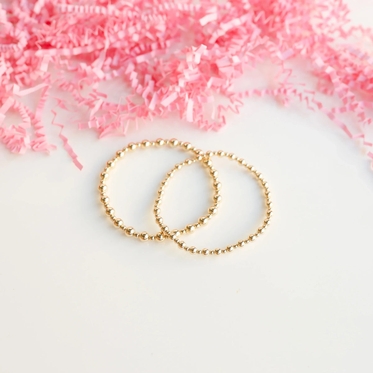 Two gold beaded bracelets arranged together against a pale pink background.