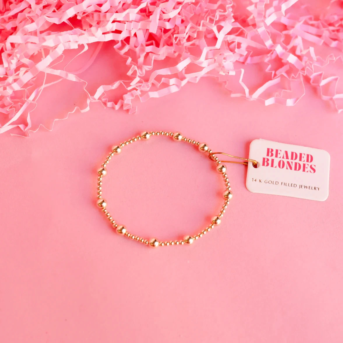 Beaded Blondes Lively Bracelet in Gold arranged with a brand tag on a pink background.