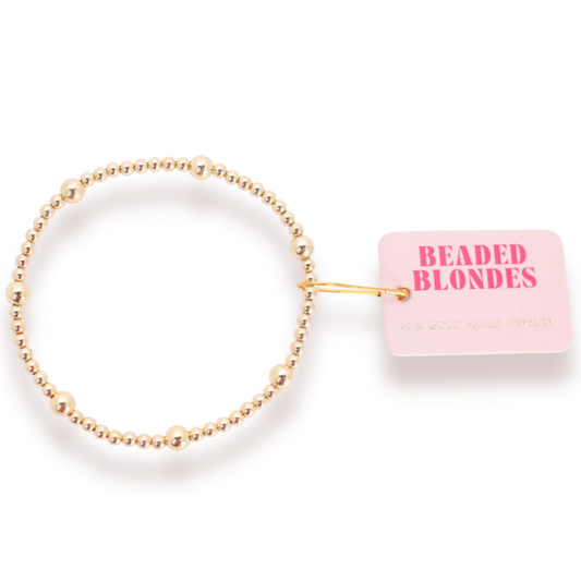 Beaded Blondes Olive Bracelet in Gold arranged in a circle, showing mixed bead sizes beside a pink tag.