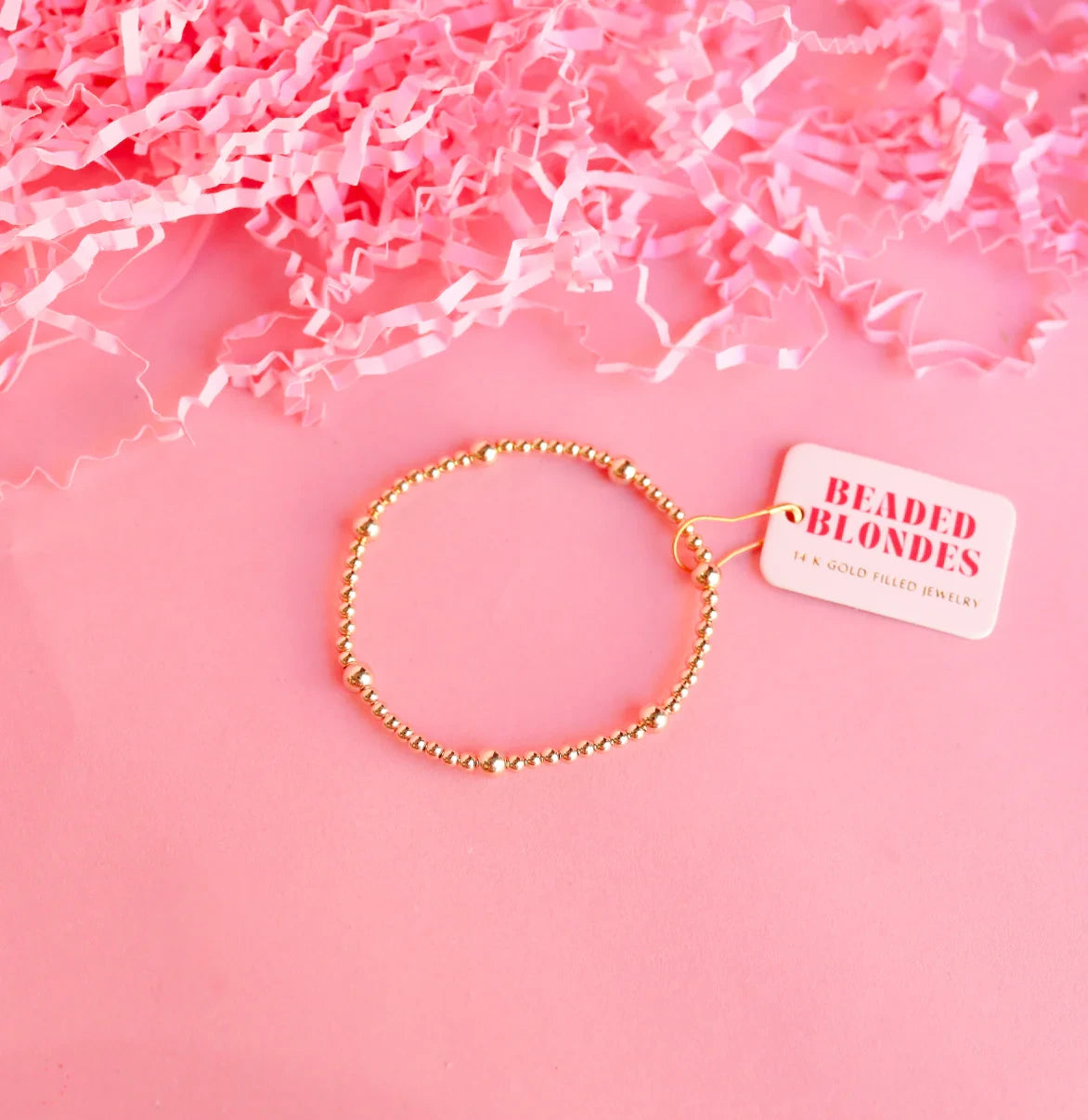 Beaded Blondes Olive Bracelet in Gold displayed on a pink surface beneath pink paper filler.