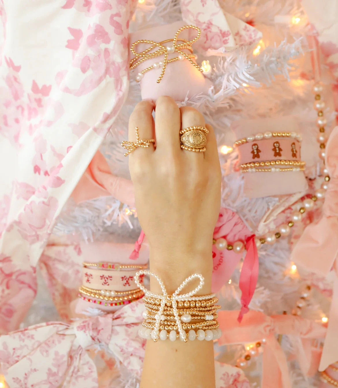 Pearl bow bracelet layered with gold beaded bracelets on a wrist beside a pink floral outfit.
