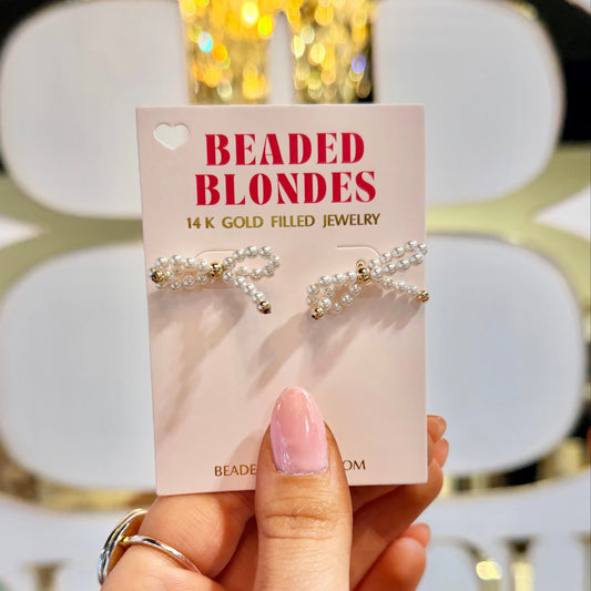 White pearl bow earrings on a Beaded Blondes card held in front of a boutique display.