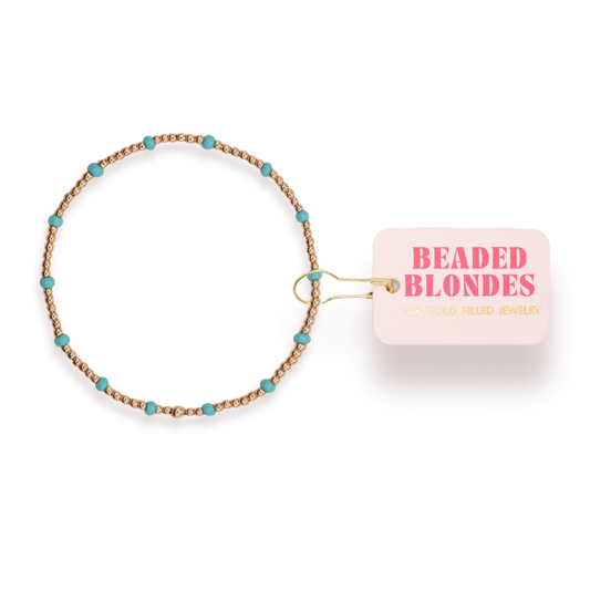 Beaded Blondes Turquoise Dainty Poppi Bracelet with turquoise-color glass accents and gold beads beside a pink tag.