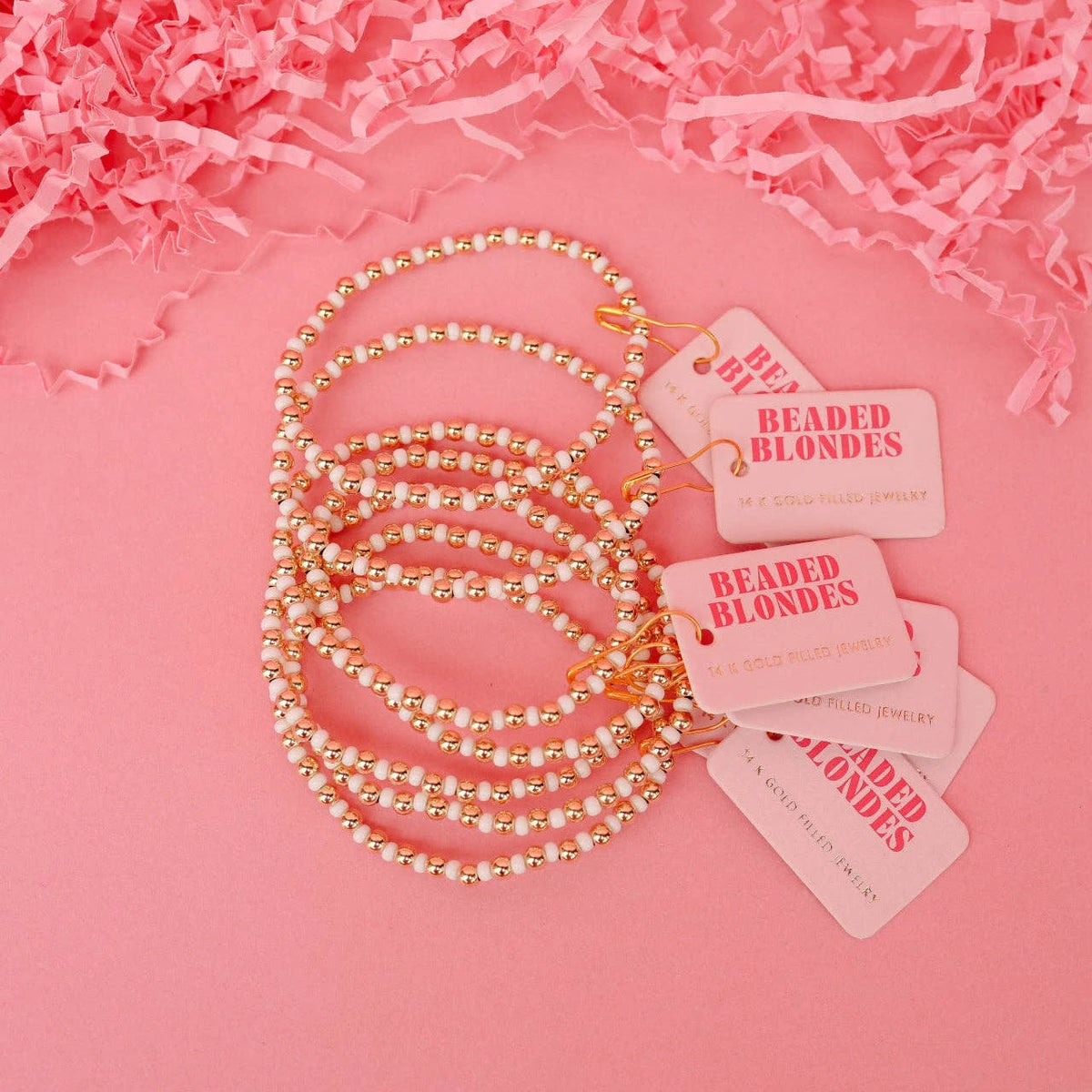Several white and gold beaded bracelets arranged with pink Beaded Blondes tags on a pink surface.