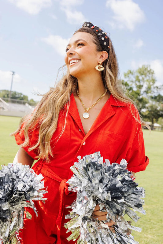 A woman outdoors on a grassy field wearing a bright red collared shirt and holding silver and black pom-poms. She is smiling and looking upward under a blue sky.