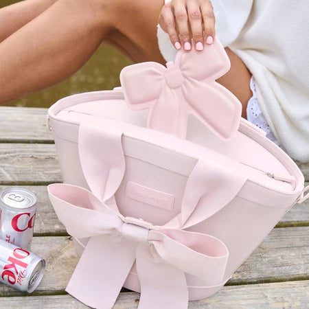 A CASE-MATE pale pink jelly ice pack with bow details, shown on a wooden table with a person's hand resting on the handle and part of their legs visible. Two beverage cans sit beside the bag.