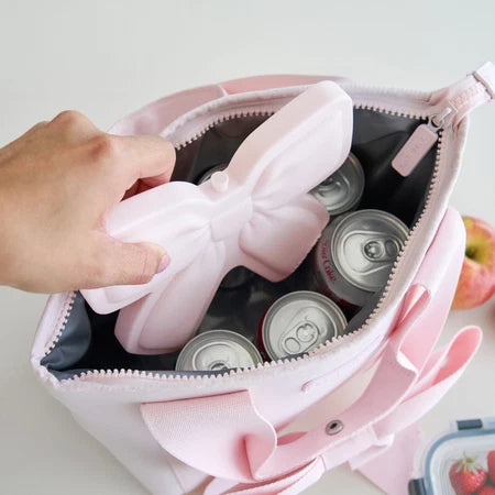 A hand placing the CASE-MATE light pink silicone bow-shaped ice pack into an open light pink cooler bag filled with canned drinks, on a white background.