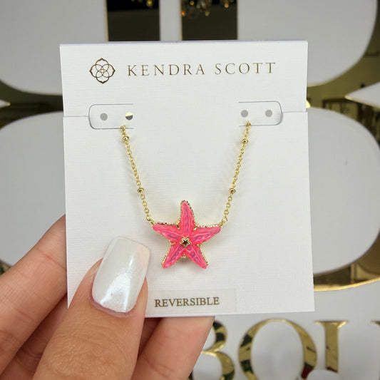 Hand holding a Kendra Scott reversible necklace with a pink starfish charm against a decorative background.
