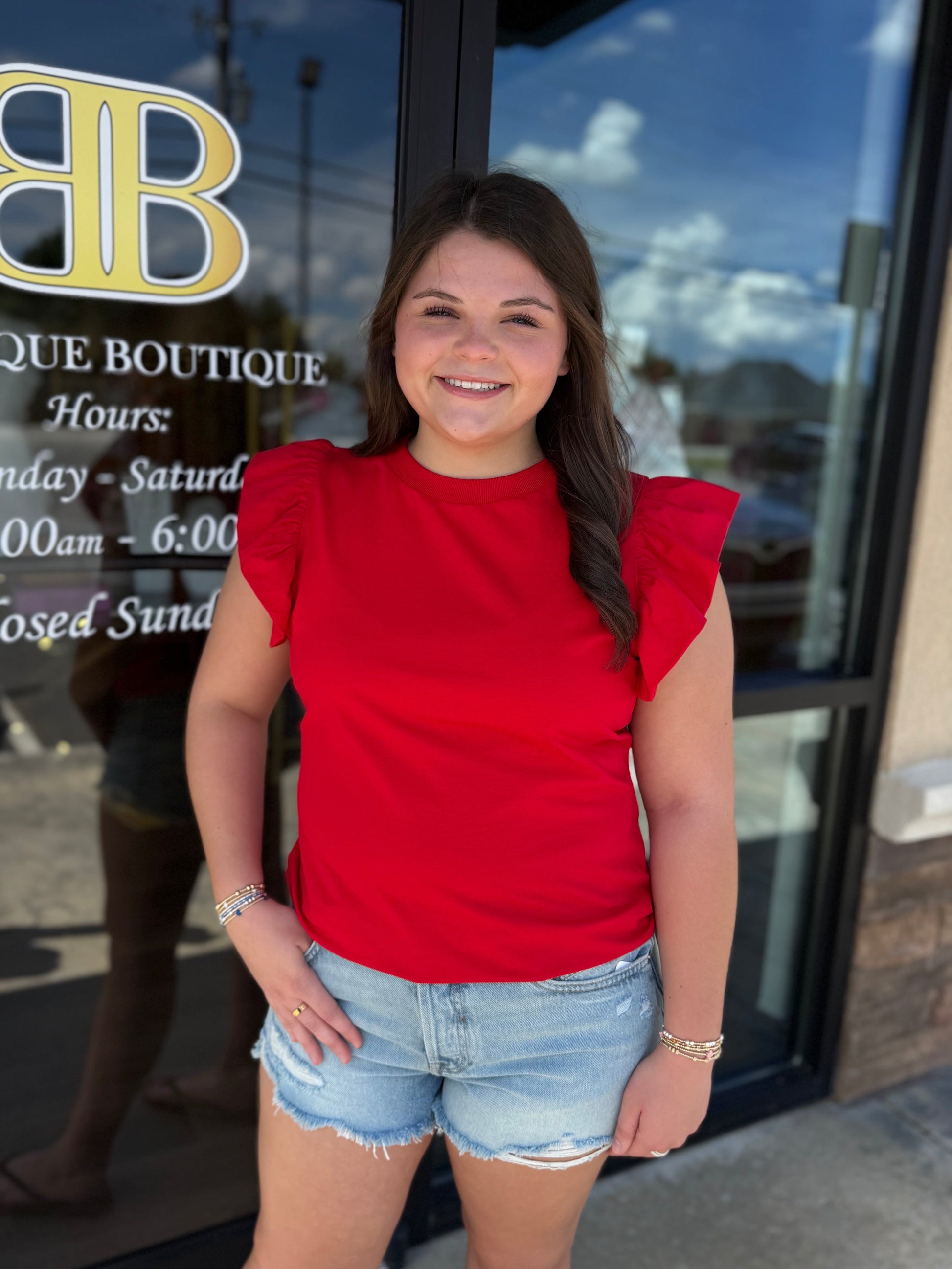 A smiling woman stands in front of a boutique glass door wearing a bright red ruffled-sleeve top and light blue denim shorts. The door has a gold and white 'BB Boutique Boutique' logo and business hours printed on it.
