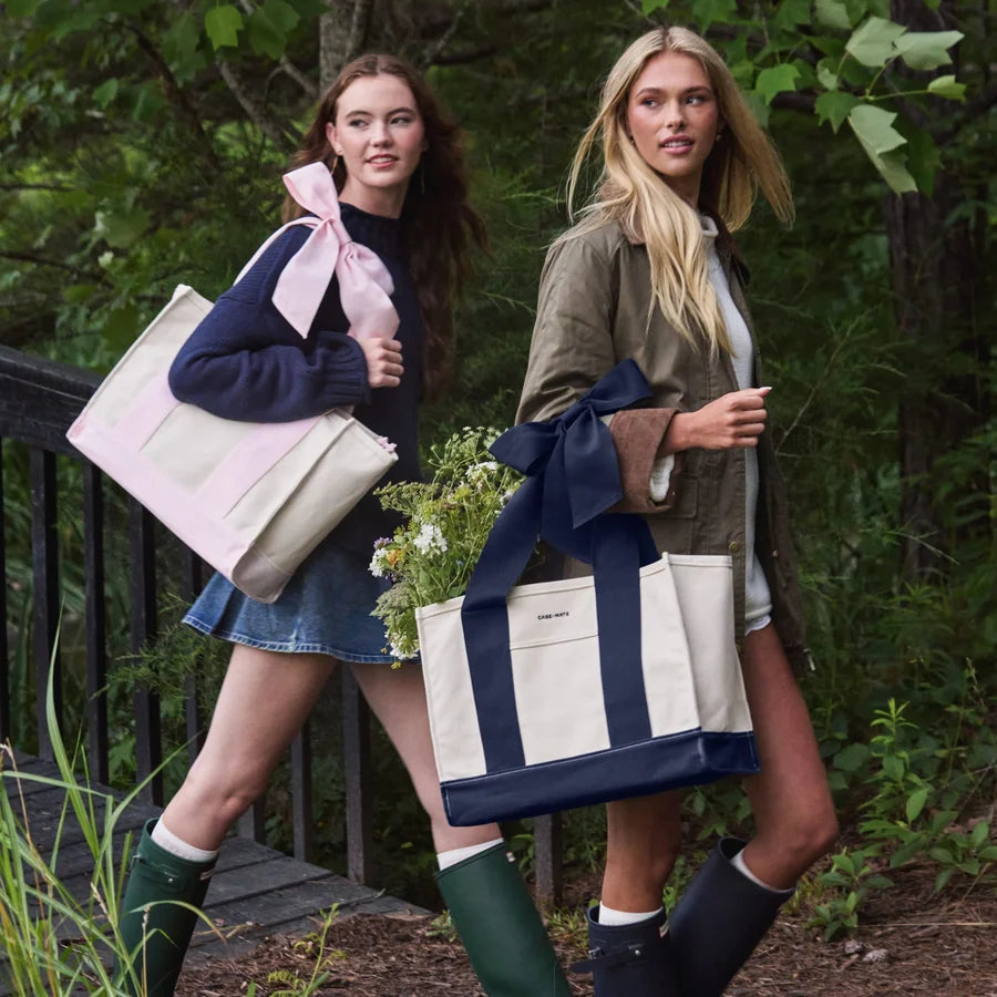 Two women walking outdoors in a wooded setting, each carrying a large canvas tote bag with a wide ribbon bow. The tote bags are cream with navy blue panels and handles.