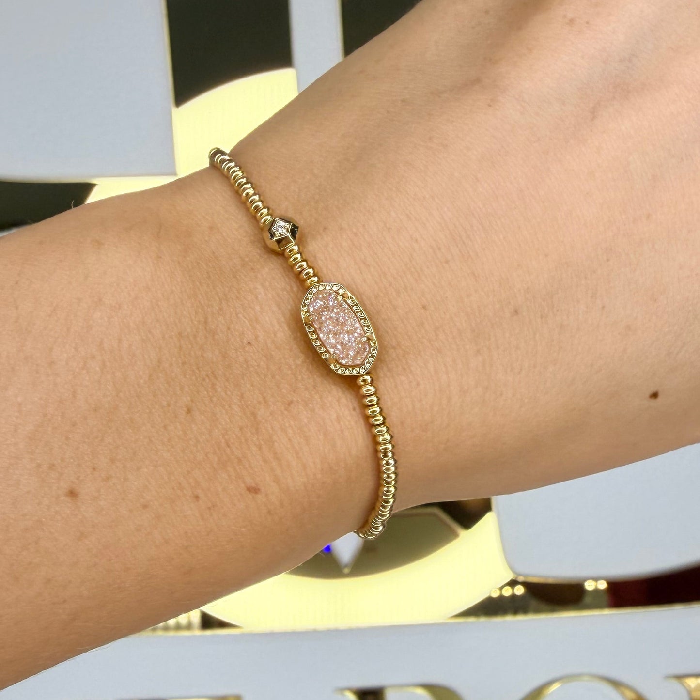 Gold bracelet on a wrist with a blurred background featuring decorative elements.