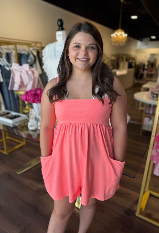 A smiling woman wearing a coral pink sleeveless romper with side pockets, standing inside a boutique store.