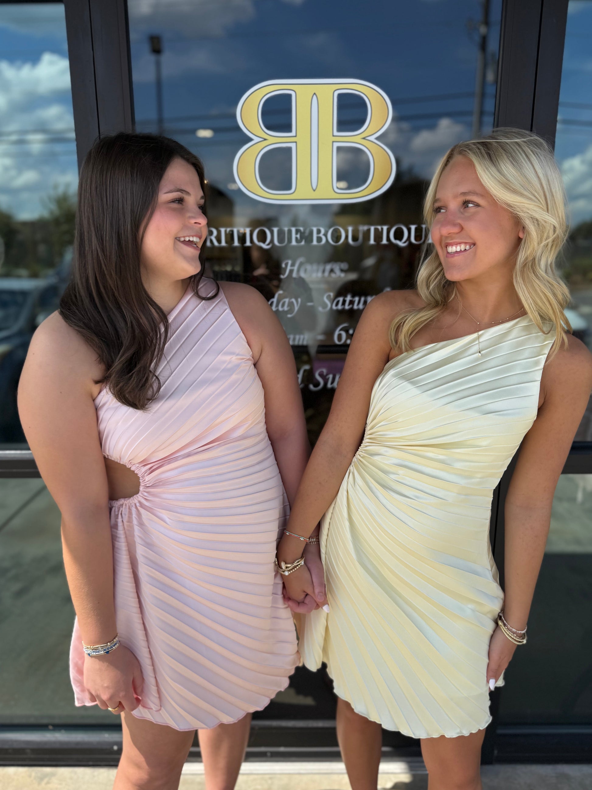 Two women standing side-by-side in front of a boutique glass door, wearing one light pink pleated dress and one cream pleated dress. The boutique door features a gold 'BB' logo and the text 'BRITIQUE BOUTIQUE'.