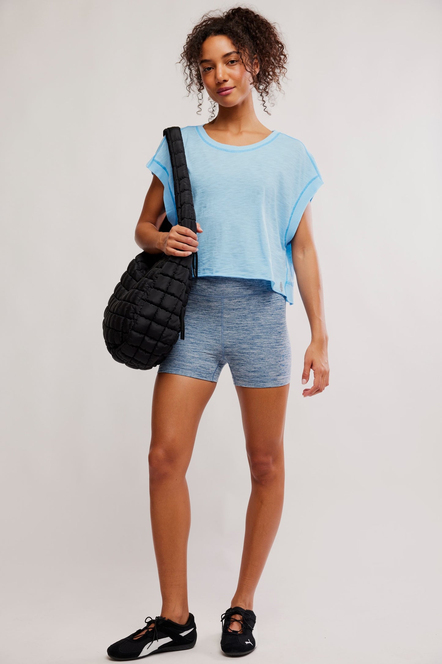 Model wearing a blue shirt with grey biker shorts holding a black bag and black shoes backing forwards
