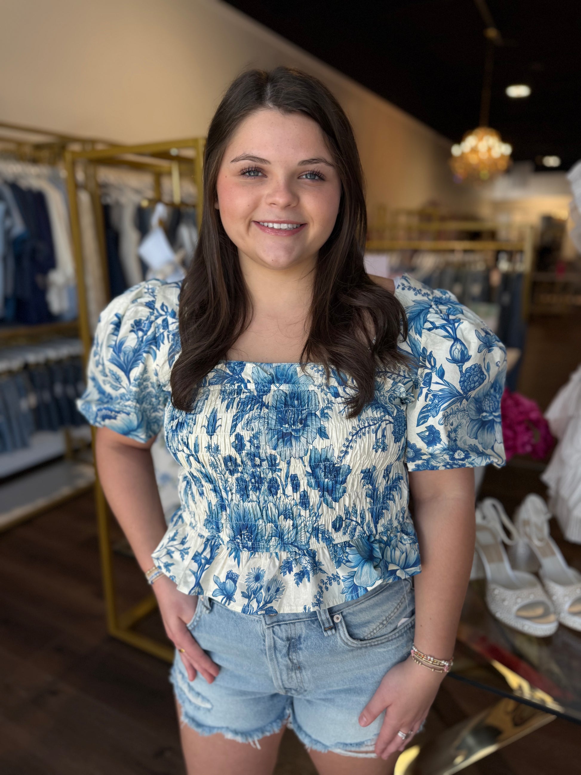 A young woman with long brown hair stands in a clothing store wearing a blue and white floral print puff-sleeve top and light-wash denim shorts.