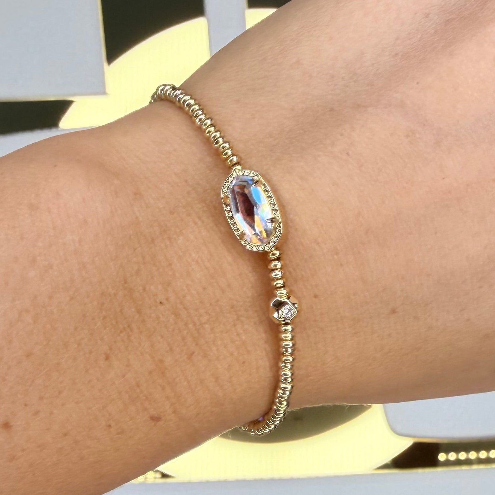 Gold Kendra Scott stretch bracelet on a wrist with a decorative background