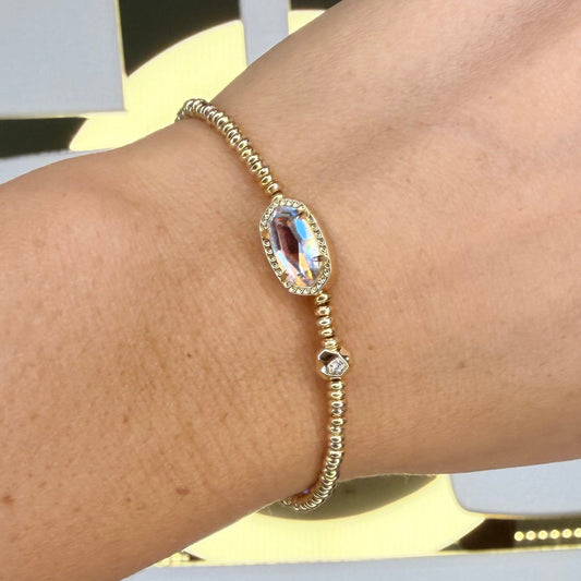 Gold Kendra Scott stretch bracelet on a wrist with a decorative background
