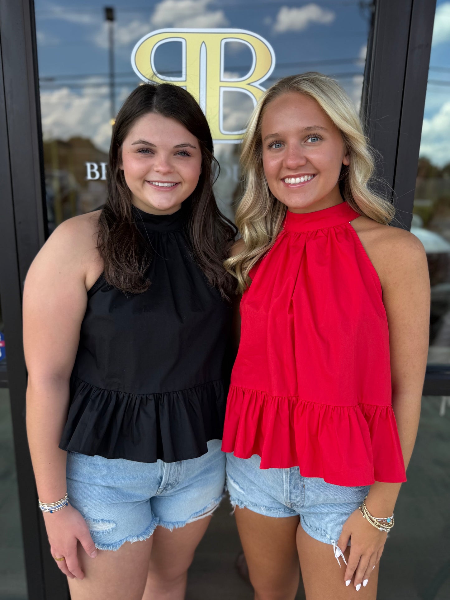 Two young women smiling and posing together in front of a glass storefront door. The woman on the left wears a sleeveless black ruffled top and light-wash denim shorts, while the woman on the right wears a bright red ruffled halter top and matching denim shorts.