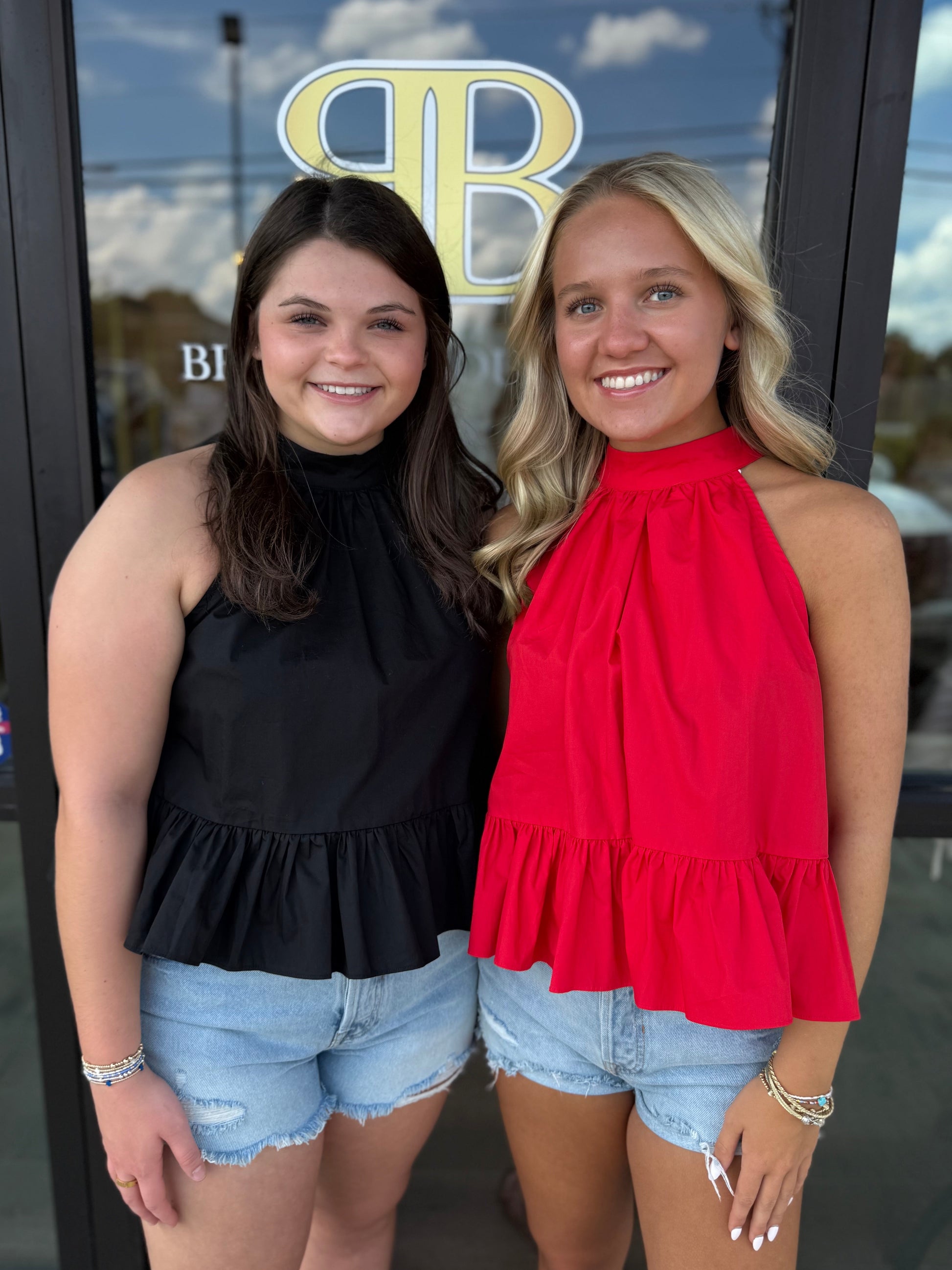 Two young women smiling and posing together in front of a glass storefront door. The woman on the left wears a sleeveless black ruffled top and light-wash denim shorts, while the woman on the right wears a bright red ruffled halter top and matching denim shorts.