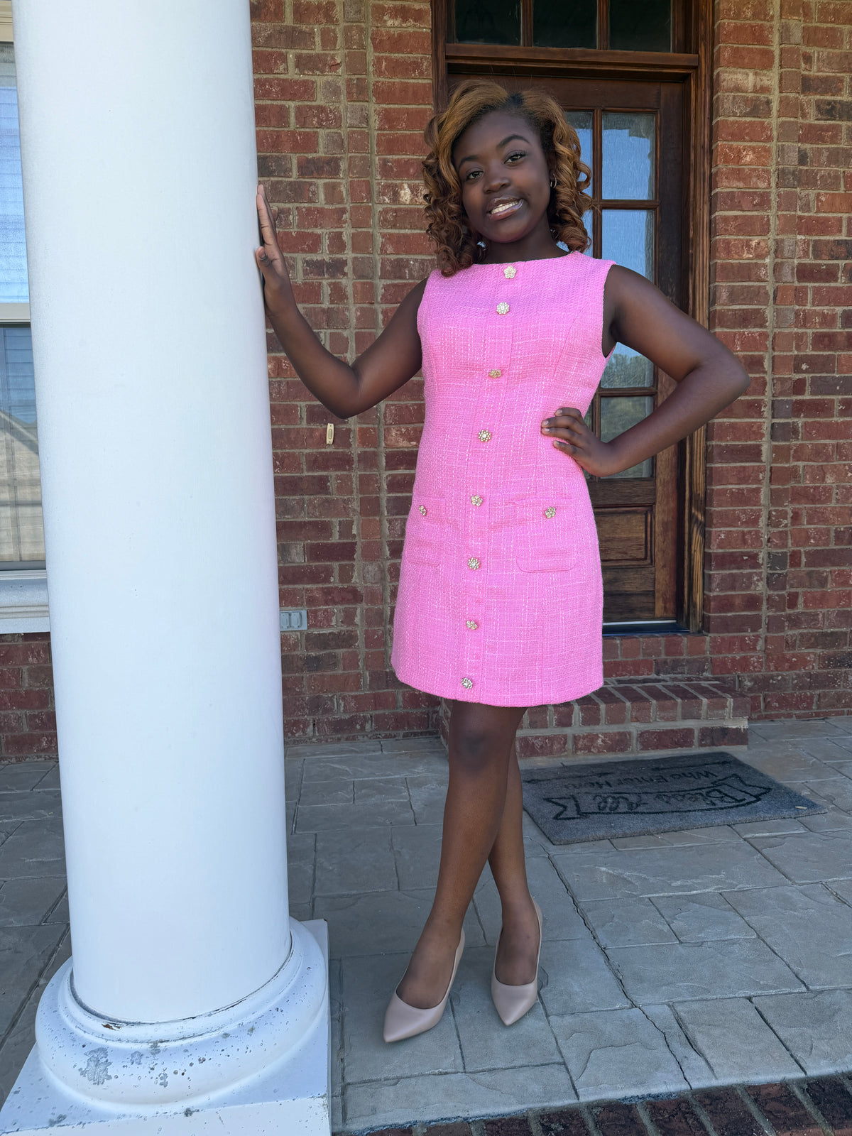 A woman stands on a brick porch wearing a bright pink sleeveless textured dress with gold buttons down the front, paired with beige heels, posing next to a white column.