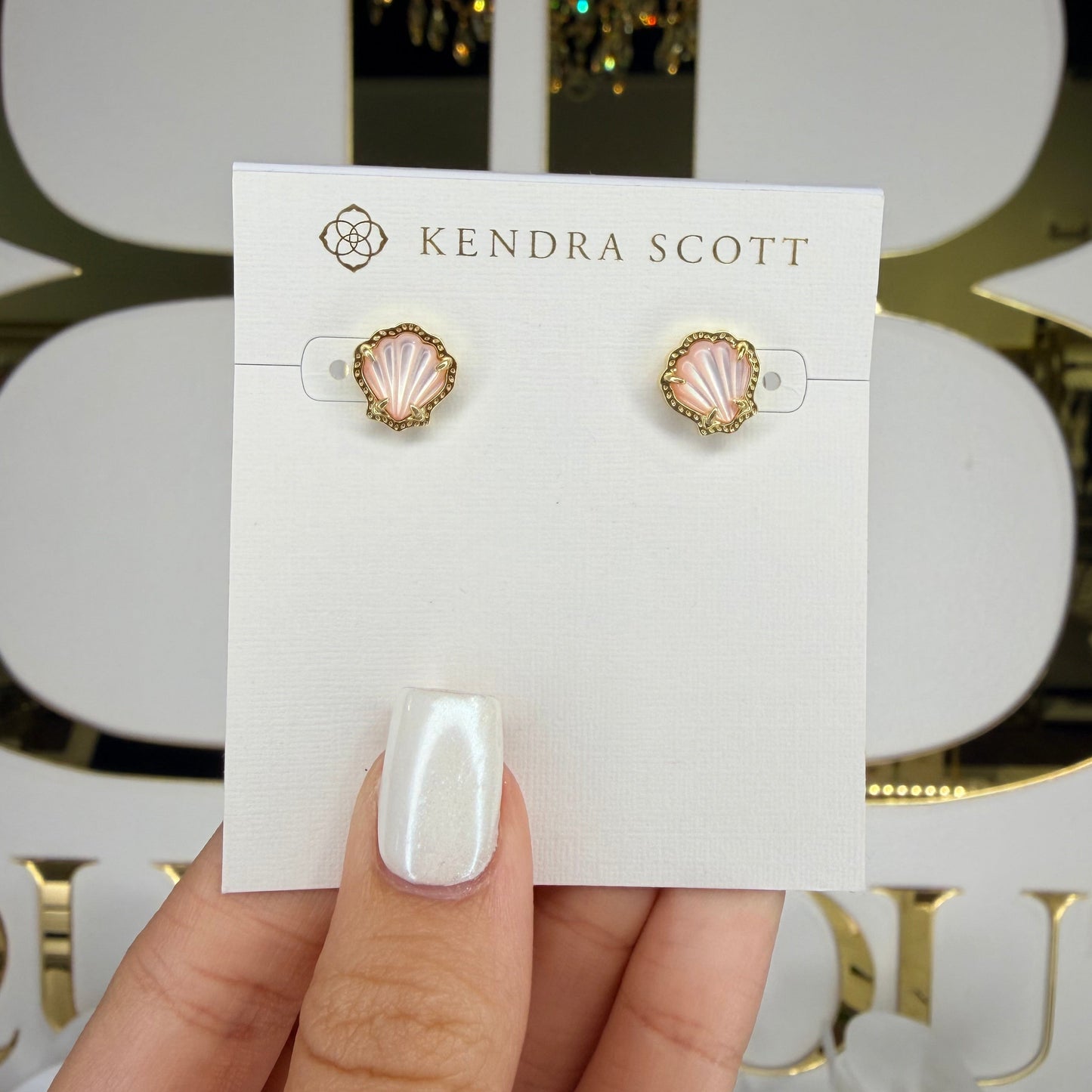Hand holding a pair of earrings on a Kendra Scott card with a decorative background.