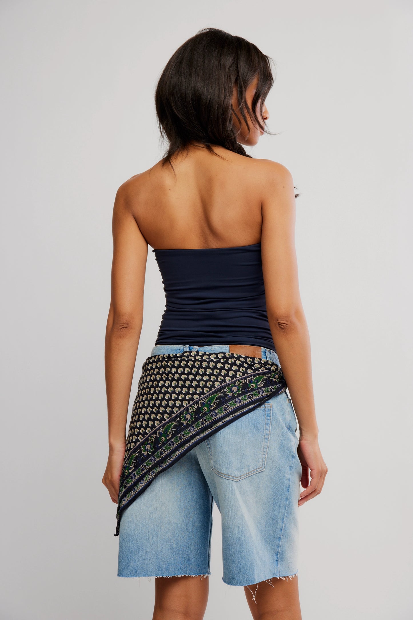 Woman wearing a strapless top and denim shorts with a patterned wrap on a plain background