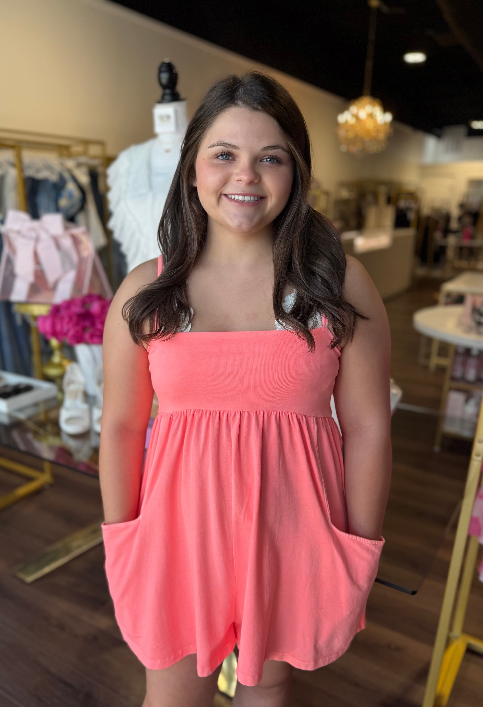 A smiling woman wearing a coral pink sleeveless romper with side pockets, standing inside a boutique store.