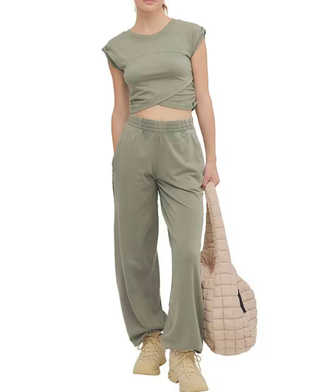 A model wearing a matching sage green two-piece set consisting of a short-sleeve wrap-style crop top and wide-leg sweatpants, paired with beige sneakers and holding a beige quilted shoulder bag against a plain white background.