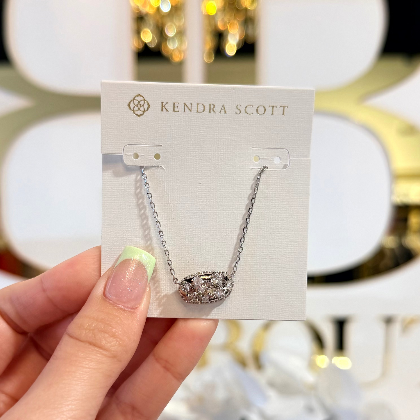 Hand holding a Kendra Scott necklace card with a blurred decorative background