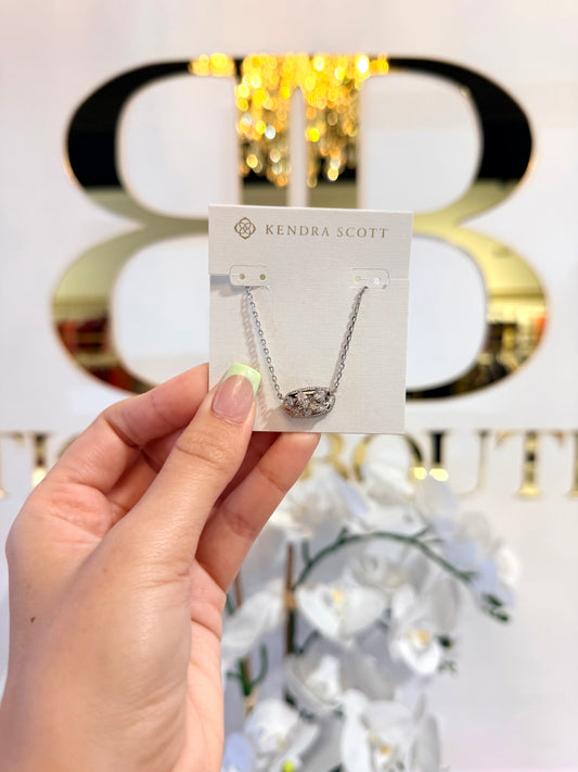 Hand holding a Kendra Scott necklace card with a blurred decorative background