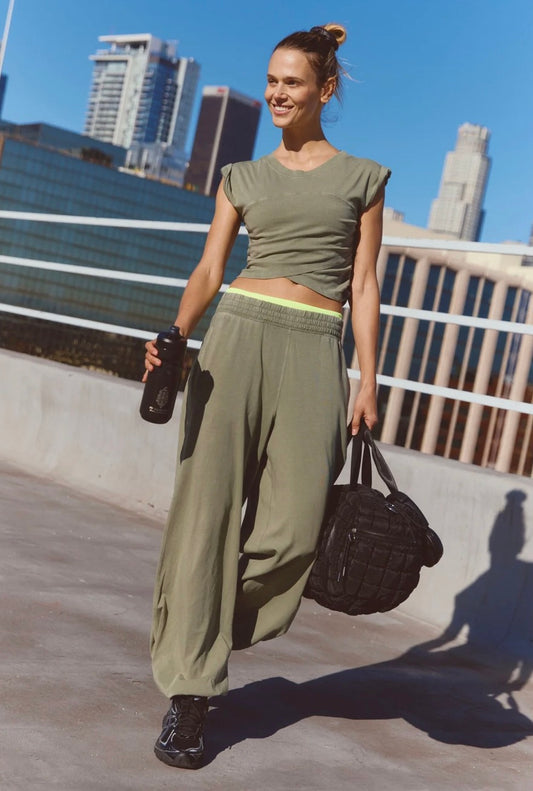 A woman walking outdoors on a rooftop wearing an olive green athletic crop top and matching wide-leg sweatpants, holding a black water bottle and carrying a black duffel bag, with a city skyline and blue sky in the background.