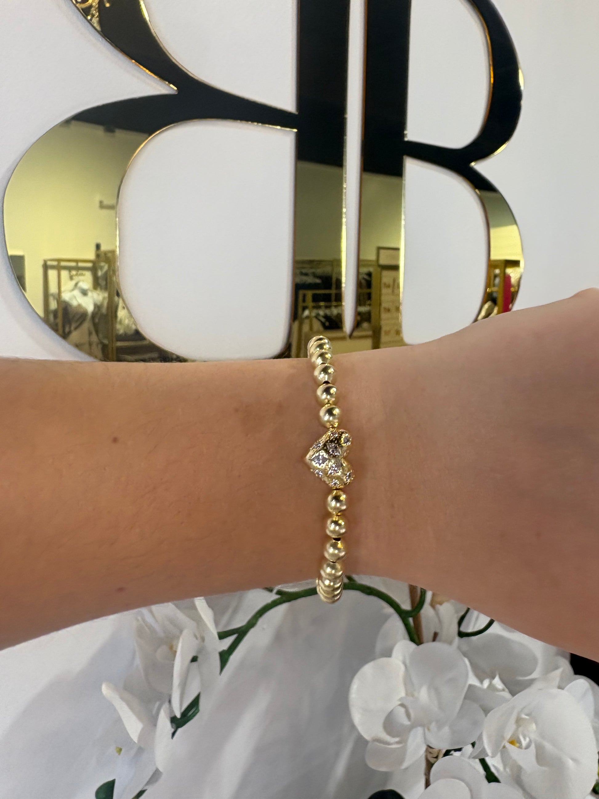 Gold KENDRA SCOTT bracelet with a heart pendant on a wrist with a blurred background.