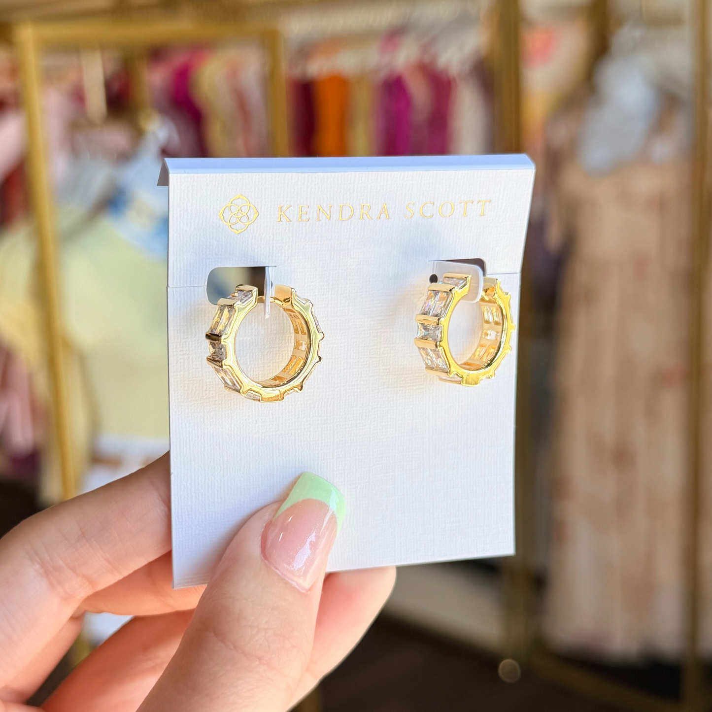 Gold hoop earrings on a Kendra Scott card held by a hand with a blurred store background.
